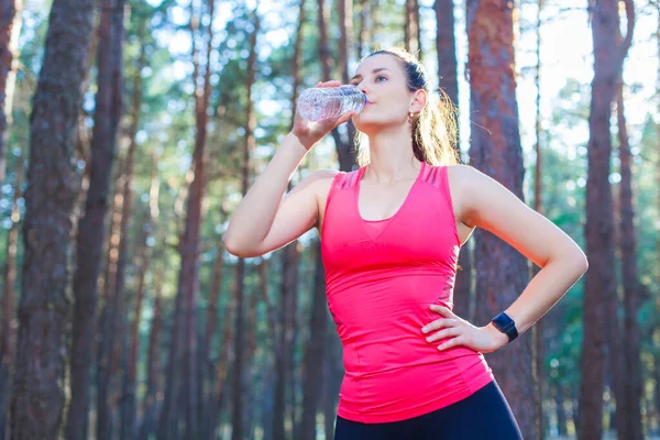 Fit λεπτή νεαρή γυναίκα σε αθλητικά ρούχα πόσιμο εμφιαλωμένο νερό, όπως η ίδια σταματά μια προπόνηση τρέχει στο δάσος — Φωτογραφία Αρχείου