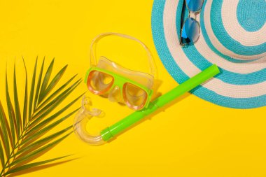 Sarı arka plandaki plaj aksesuarları. Güneş gözlükleri, çizgili mavi şapka, dalış maskesi ve palmiye yaprağı. Uzun zamandır beklenen deniz ve seyahat tatili kavramı. Reklam alanı
