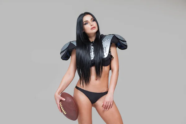 Beautiful sexy brunette female american football player in uniform posing with a ball isolated on grey background — Stock Photo, Image