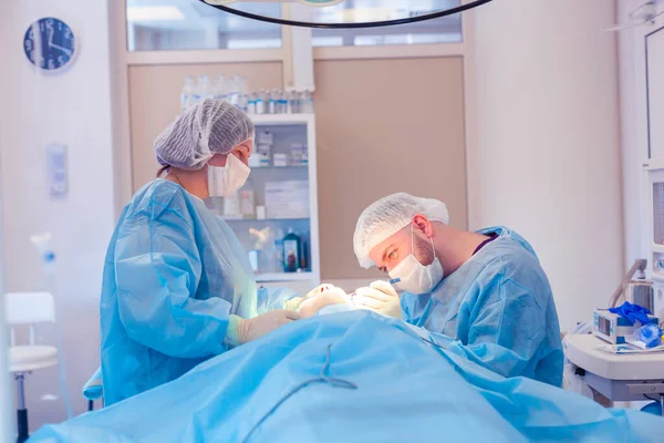 Cirujano masculino realiza procedimientos quirúrgicos en el quirófano, mientras que su asistente supervisa la condición del paciente —  Fotos de Stock