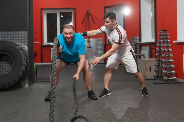 O jovem gordo está muito concentrado realizando exercícios de corda de batalha no ginásio de fitness sob controle de seu treinador pessoal. Peso em excesso — Fotografia de Stock
