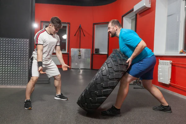 Hombre gordo es muy persistente haciendo ejercicio de volteo de neumáticos en el gimnasio bajo la guía de entrenador personal. Sobrepeso — Foto de Stock