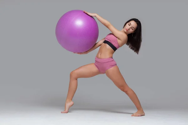 Portrait pleine longueur de fille sportive en tenue de sport rose posant avec balle de fitness au-dessus de sa tête isolée sur fond gris — Photo