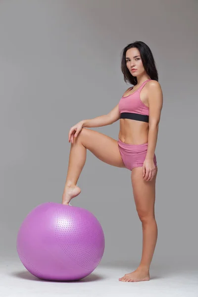 Menina atraente nova no sportswear rosa moderno está posando colocando seu pé na bola de fitness isolado no fundo cinza. Comprimento total — Fotografia de Stock