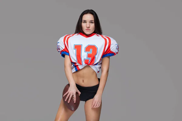 Joven hermosa morena vistiendo el uniforme sexy de jugador de fútbol americano posando con la pelota con la mano en las caderas aisladas sobre fondo gris — Foto de Stock