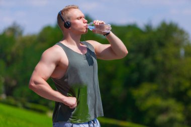 Kulaklıklı kaslı genç adam su içiyor ve yaz parkında koştuktan sonra dinleniyor.