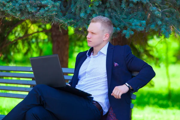 Handsome businessman in blue suit sitting on the bench in summer park working remotely on a laptop.