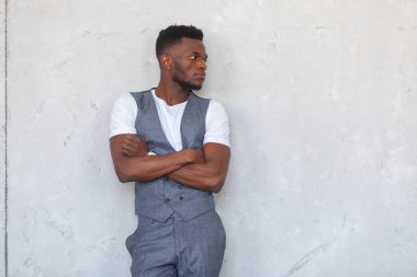 Gri yelek ve pantolonla gri beton bir duvara karşı çapraz kollu poz veren Afrikalı genç yakışıklı bir zenci.