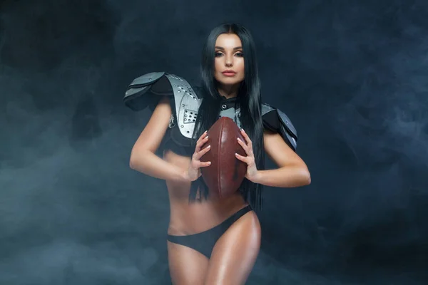 American football. Young sporty brunette wearing sexy uniform of rugby football player posing with ball in smoke isolated on black background — Stock Photo, Image