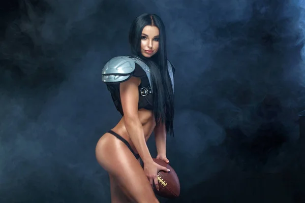 Football americano. Giovane bruna sportiva indossa sexy uniforme di giocatore di rugby calcio in posa con palla in fumo isolato su sfondo nero — Foto Stock