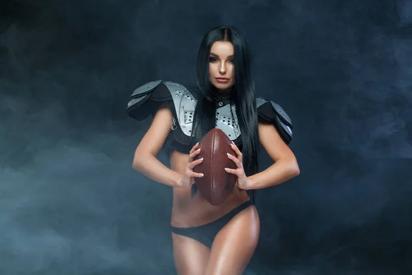 Futbol amerykański. młody sportowe brunetka noszenie sexy mundur z rugby piłkarz pozowanie z piłka w dym odizolowany na czarny tło — Zdjęcie stockowe