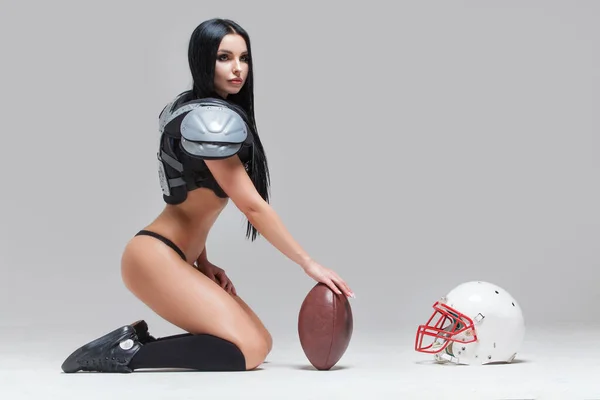 Vista lateral de la hermosa morena sexy vestida con bikini y uniformes de fútbol americano posando sentada en el suelo con una pelota y casco aislados sobre fondo gris — Foto de Stock
