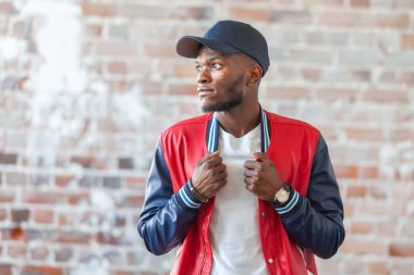 Sıradan giyinmiş yakışıklı Afrikalı bir adam harap olmuş kırmızı tuğla duvarın arka planında duruyor.