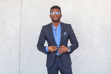 Kendinden emin Afrikalı genç bir iş adamı modern takım elbise ve mavi gömlekle gri beton bir duvara bakıyor.