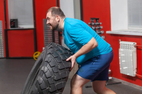 胖男人在私人教练的指导下，坚持在健身房做轮胎翻转运动。体重超标 — 图库照片