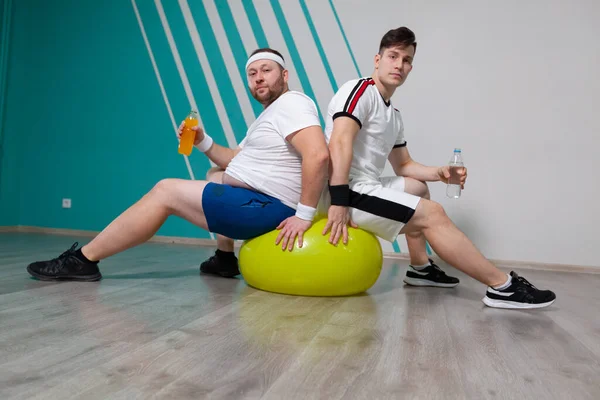 胖男人和他的私人教练在健身班的艰苦训练后，坐在健身球上，拿着瓶装水。那个超重的男人非常疲倦 — 图库照片
