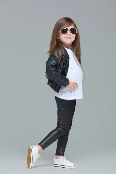 Baby girl with long hair in black leather jacket and leggings is posing in the studio in modern sunglasses on grey background