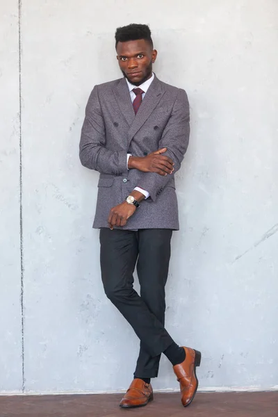 Teljes hosszúságú portré egy jóképű afrikai üzletemberről, aki modern szürke öltönyben pózol egy szürke betonfalnak támasztva. — Stock Fotó