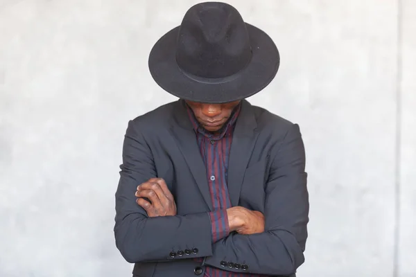 Fiatal, komoly afrikai férfi fekete öltönyben elrejti arcát egy kalap alatt, keresztbe tett karokkal áll egy szürke betonfalnak támasztva. — Stock Fotó