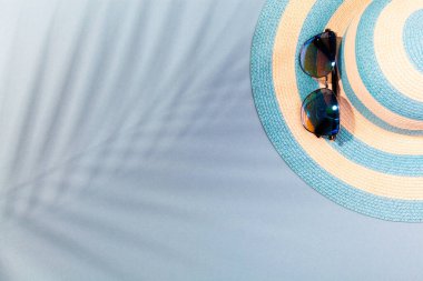 Turkuaz mavi arka planda plaj aksesuarları. Güneş gözlükleri ve palmiye yaprağının gölgesinde çizgili mavi şapka. Plaj tatili kavramı, deniz turu, sıcak güneşli yaz. Reklam alanı