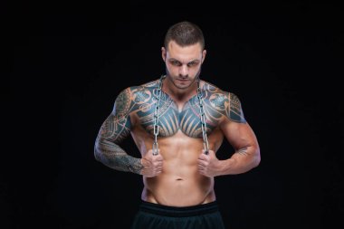 Dövmeli güçlü kaslı atletik adam siyah arka planda boynuna metal zincir takıyor.