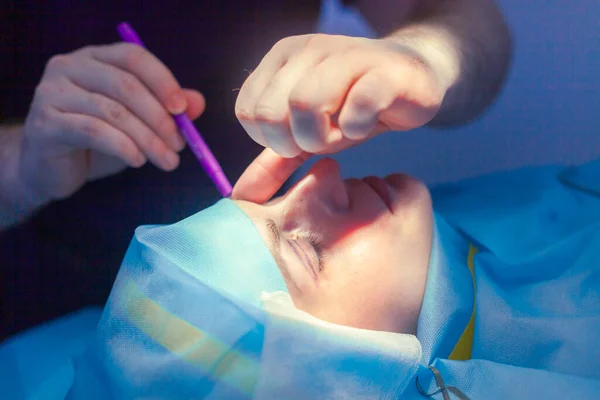 Zbliżenie męskich rąk chirurga wykonujących zabiegi chirurgiczne na twarzy pacjenta przed operacją blepharoplastyki — Zdjęcie stockowe