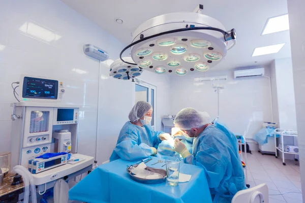 Foto de un monitor de signos vitales y equipo médico junto a dos cirujanos que realizan cirugía en el quirófano —  Fotos de Stock