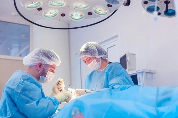 Cirujano masculino realiza procedimientos quirúrgicos en el quirófano, mientras que su asistente supervisa la condición del paciente —  Fotos de Stock
