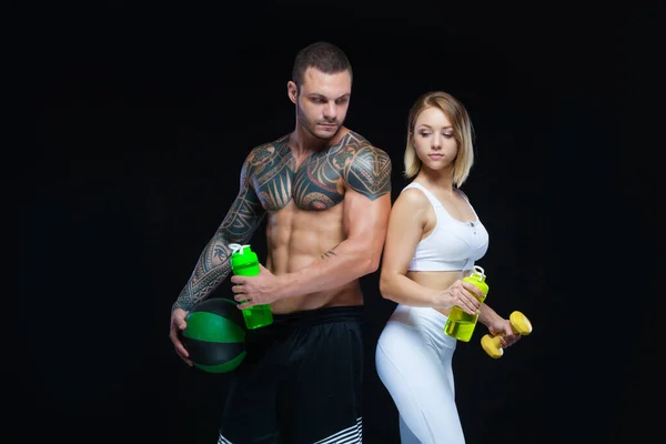 Casal de fitness - mulher e homem muscular tatuado posando com garrafas de água e equipamentos esportivos sobre fundo preto — Fotografia de Stock