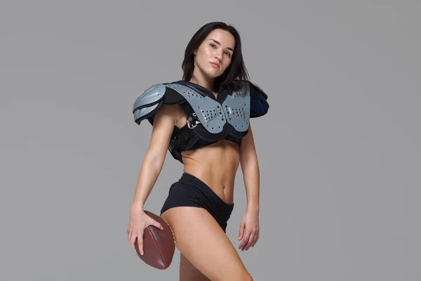 Junge sportliche Brünette in sexy Uniform eines American-Football-Spielers posiert mit Ball und blickt isoliert in die Kamera auf grauem Hintergrund — Stockfoto