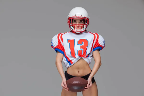 Sportliches Mädchen in sexy Uniform des American Football-Spielers und Helm posiert mit Ball bereit, isoliert auf grauem Hintergrund zu werfen — Stockfoto