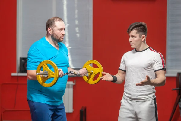 Um homem gordo no ginásio realiza exercícios de levantamento de sinos no limite de seus poderes sob a orientação de um personal trainer. Sobrepeso — Fotografia de Stock