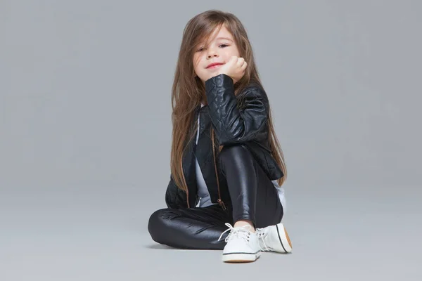 Niña con el pelo largo en chaqueta de cuero negro y leggings está sentado en el estudio sobre fondo gris — Foto de Stock