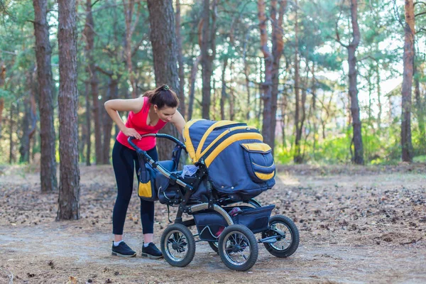 Nature walk with stroller, young active mother in sportswear walking on the forest walkway with her baby in the pram, enjoying fresh air and smiling to the child — Stock Photo, Image
