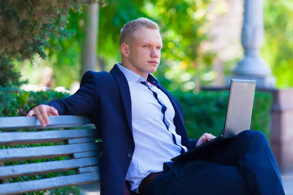 Handsome businessman in blue suit sitting on the bench in summer park working remotely on a laptop.