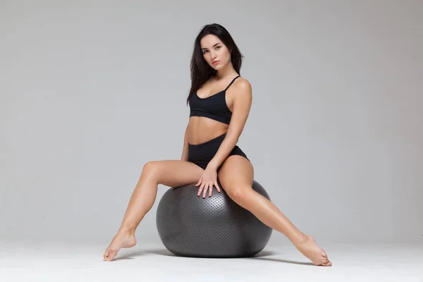 Full length portret van sportieve sexy meisje zitten op fitness bal stretching haar been uit geïsoleerd op grijze achtergrond — Stockfoto