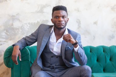 Tarz sahibi Afrikalı siyahi bir adam stüdyoda yeşil bir kanepede oturuyor. Gri beton bir duvara karşı üç parça takım elbise içinde.