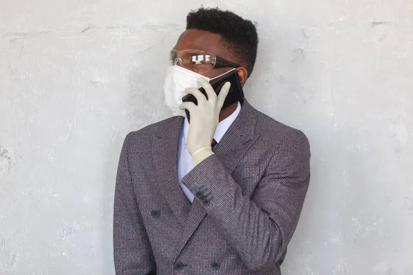 COVID-19. Takım elbiseli yakışıklı Afrikalı siyahi adam tıbbi maske takıyor, koruyucu gözlük takıyor ve elinde eldivenle cep telefonunu tutuyor. Koronavirüs enfeksiyonunu önlemek için önlemler. — Stok fotoğraf