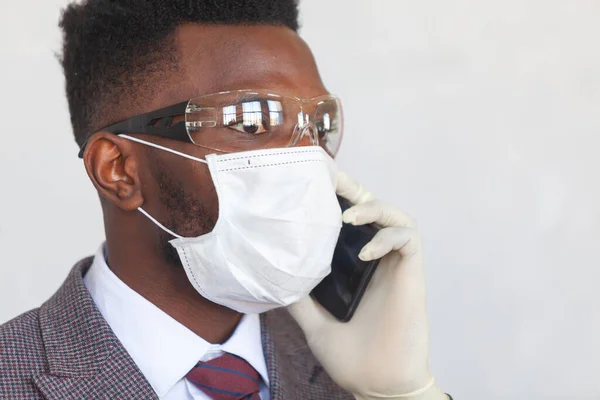 COVID-19. Красивый африканский черный мужчина в костюме использует медицинскую маску, защитные очки и держит свой мобильный телефон рукой в перчатках. Мясо для предотвращения коронавирусной инфекции и — стоковое фото