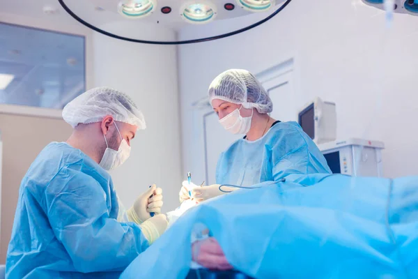 Cirurgião masculino realiza procedimentos cirúrgicos na sala de cirurgia, enquanto seu assistente monitora a condição do paciente — Fotografia de Stock