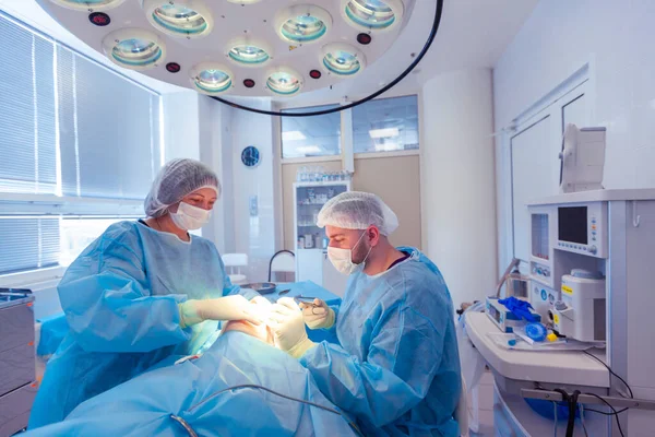 Cirurgião masculino realiza procedimentos cirúrgicos na sala de cirurgia, enquanto seu assistente monitora a condição do paciente — Fotografia de Stock