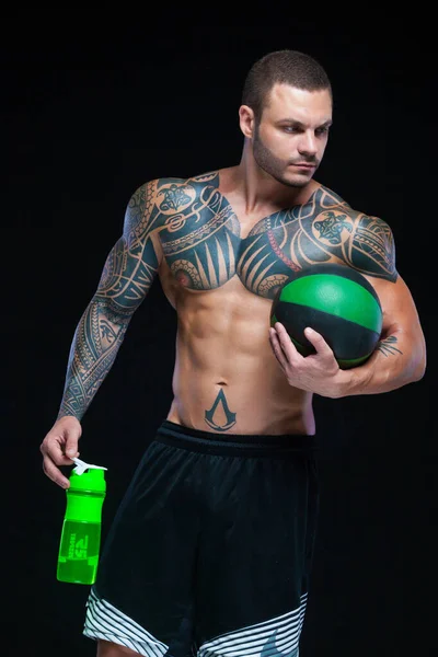 М'язовий чоловік культурист з татуюваннями. Чоловік позує на чорному тлі з пляшкою води і м'ячем — стокове фото