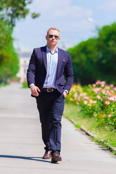 Joven hombre guapo en traje azul agradable está caminando en el parque de verano en gafas de sol — Foto de Stock