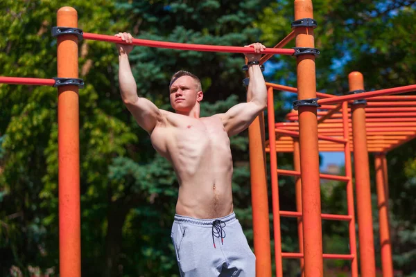 Çıplak gövdeli güçlü kaslı bir adam kendini direğin üzerine çekiyor. Yaz parkında dışarıda çalışıyorum.
