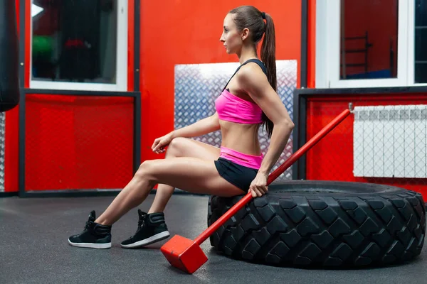 Vista lateral da aptidão muscular mulher morena está posando no ginásio com a marreta sentada no pneu — Fotografia de Stock