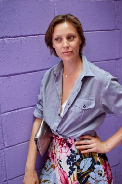 Mor tuğla duvarın arkasında poz veren güzel bir kız. O şık bir yatak ovetvet gömlek ve uzun parlak bir etek giyiyor..