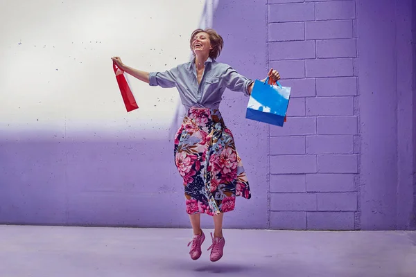 Joven chica hermosa saltando de alegría con compras en sus manos. Una chica con una falda larga y brillante y un top rosa pálido está caminando desde la tienda . — Foto de Stock