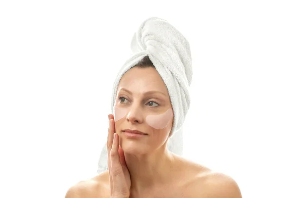 Piękna kobieta w średnim wieku z ręcznikiem na głowie z łatami na oczy przed zmarszczkami. Białe pojedyncze tło. — Zdjęcie stockowe