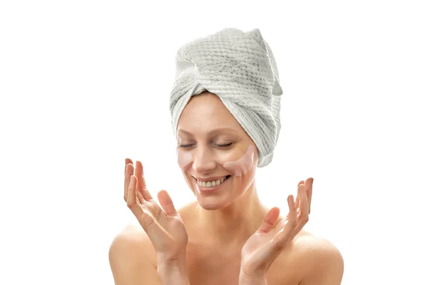 Piękna kobieta uśmiechnięta z ręcznikiem na głowie z łatami na oczy przed zmarszczkami. Białe pojedyncze tło. — Zdjęcie stockowe