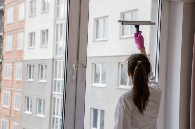 Güzel genç bir kız pembe eldivenlerle pencereleri temizlemek için özel bir fırçayla pencereyi yıkıyor. Pencerenin dışında çok katlı binalar var..